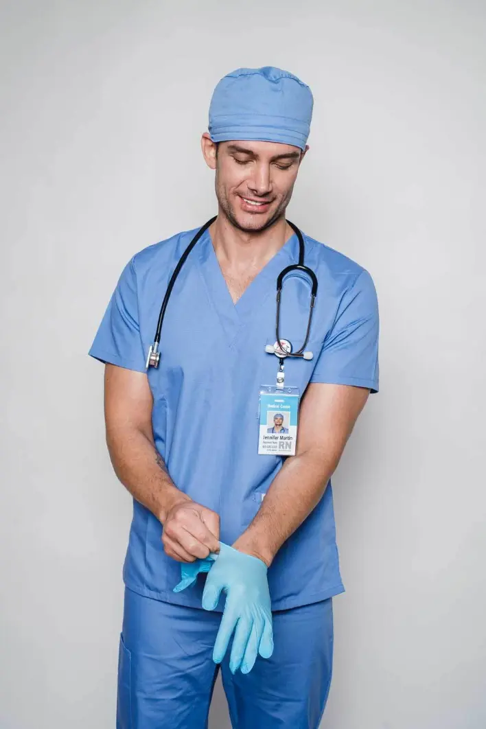 Portrait d’un professionnel de santé portant une tenue bleue de bloc opératoire, un calot et un stéthoscope autour du cou. Il enfile des gants médicaux avec un badge d’identification visible, ce qui évoque la préparation avant intervention, les gestes d’hygiène et la rigueur hospitalière. L’image est idéale pour illustrer des contenus sur les infirmiers, chirurgiens, soins hospitaliers ou protocoles sanitaires.